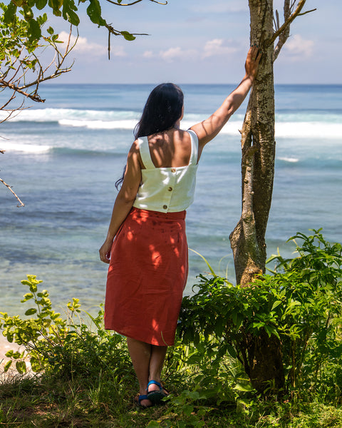 A woman in a white top and orange skirt stands with their back to the viewer, looking out at the ocean. They are leaning against a tree on a grassy bank.