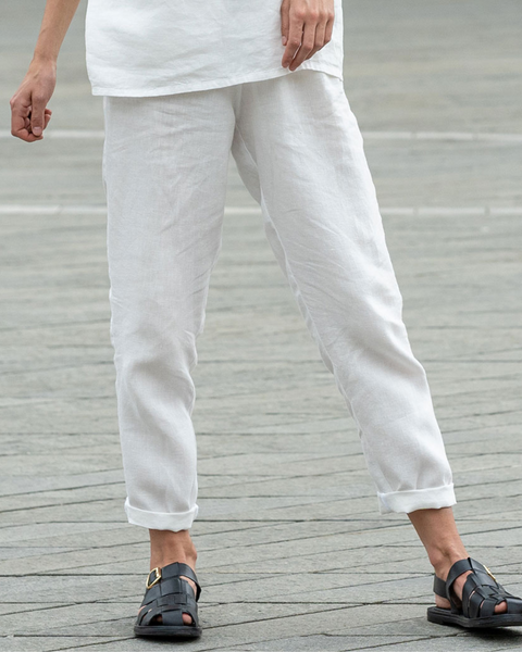 A person stands with their feet apart on a paved surface, viewed from the waist down. They are wearing casual pure white menique linen pants that are slightly wrinkled and cuffed just above the ankles. On their feet are dark, likely black, sandals with multiple straps and buckles. The paving stones are in shades of grey and beige, and the overall lighting suggests an outdoor setting.