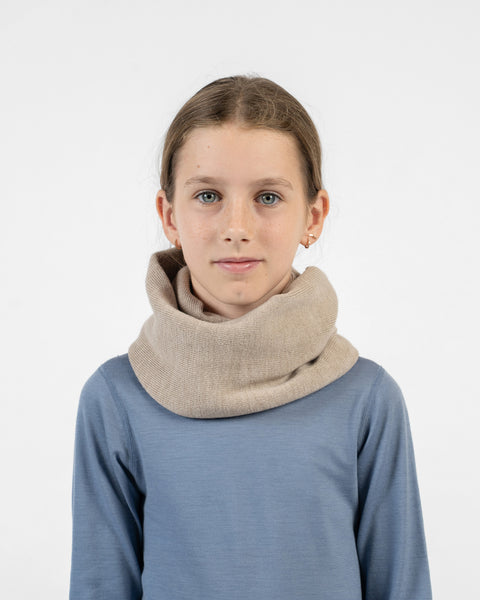 A young girl looking directly forward, wearing a beige knit infinity scarf around her neck and a blue long-sleeved shirt.