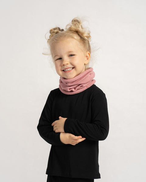 Little girl in a black outfit wearing a soft pink knit neck warmer, smiling with arms crossed.