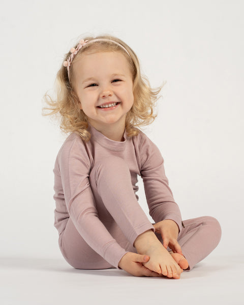 A smiling child with blonde curly hair and a pink floral headband sits cross-legged on a white background, wearing a dusty pink long-sleeved top and matching pants. The child is looking directly at the camera.