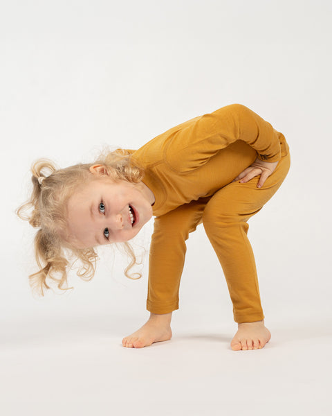 A playful child with curly blonde pigtails, wearing a yellow long-sleeved top and matching pants, is bending over with their hands on their hips, looking up at the camera with a wide smile. The child is barefoot on a white surface.