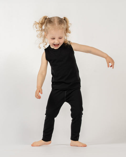 A young child with blonde, curly hair styled in pigtails, is standing barefoot against a white background. The child is wearing a black sleeveless top and black pants. They are looking down and to their left with a wide smile and open mouth, appearing to be in motion or mid-laugh. Their arms are slightly extended out from their sides.