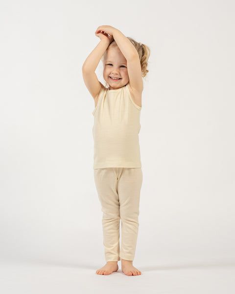 A young child with light, curly hair tied back is standing barefoot against a plain white background. They are wearing a natural colored sleeveless top and matching leggings. The child is smiling widely at the camera with their arms raised above their head, hands clasped together, in a playful pose.