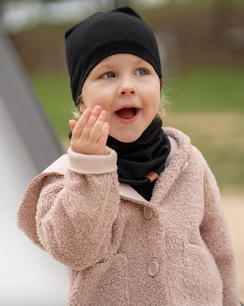 Little girl in black Merino beanie and neck gaiter blowing a kiss, dressed in a beige teddy coat, outdoor playground setting.