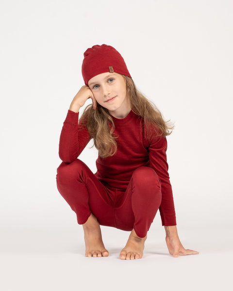 a girl, crouching down, wearing the kids' merino beanie in the color royal cherry, and a matching color merino set with long sleeve top and pants.