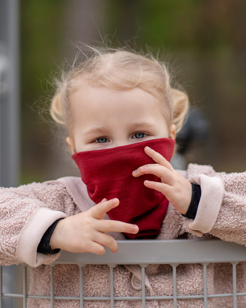 A young child with blonde hair and blue eyes looks over a railing, wearing a red Menique neck gaiter pulled up to cover their nose and mouth. Their small hands are resting on the railing, with a beige jacket visible.