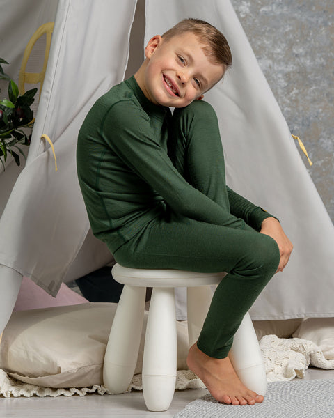 A boy sitting an a chair, with one leg on top of it, wearing the menique Kids' 160 Merino Pants in Dark Green color. He is resting his head on the raised leg's knee.