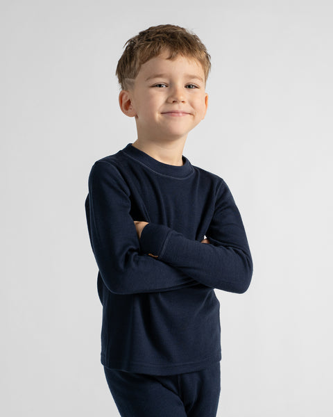 A young boy with short light brown hair stands against a light gray studio background, wearing a dark navy long-sleeve crewneck shirt and matching navy pants.