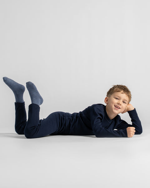 A young boy lying on his stomach on a light gray studio floor, wearing a dark navy long-sleeve pajama set and matching navy socks.