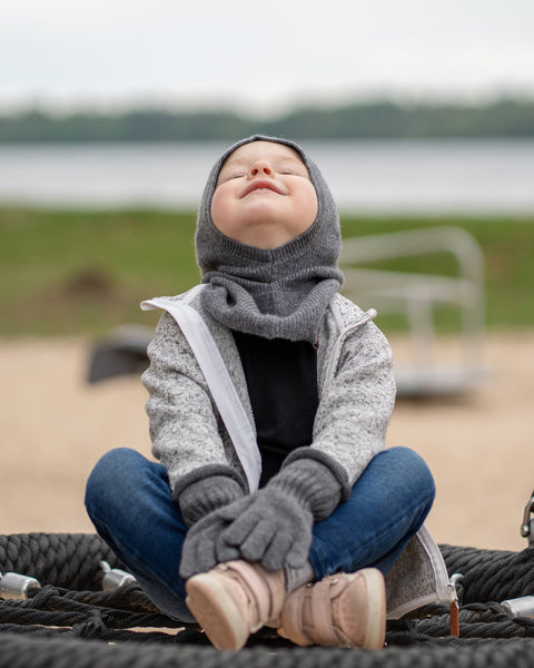 A young child sits cross-legged on a black rope swing, looking upwards with a serene smile. They are wearing a dark grey balaclava, a grey speckled zip-up hoodie, dark grey mittens, blue jeans, and light pink sneakers. The background is a blurred outdoor scene with sand, grass, and water.