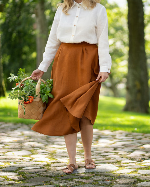 A woman strolls along a cobblestone park pathway, carrying a woven basket filled with fresh produce and green sprigs. She is wearing a crisp white linen button-down shirt and a flowing almond brown maxi skirt, playfully lifting the skirt's hemline as she walks in brown sandals.