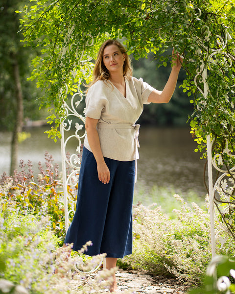 A woman stands barefoot beneath a white metal garden archway covered in lush green leaves, gazing thoughtfully to the side. She is modeling a natural-colored, short-sleeve linen wrap top with a tie waist and wide-leg, storm blue linen pants.