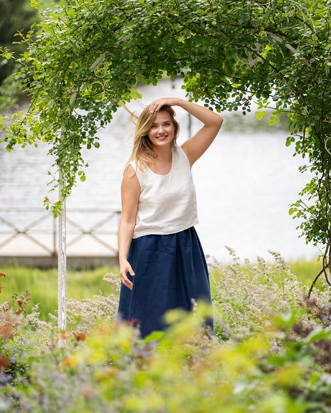 A woman smiles brightly at the camera while standing under a white decorative garden archway draped in green vines near a lake. She wears a sleeveless pure white linen top tucked into a storm blue midi skirt with a gathered waist.
