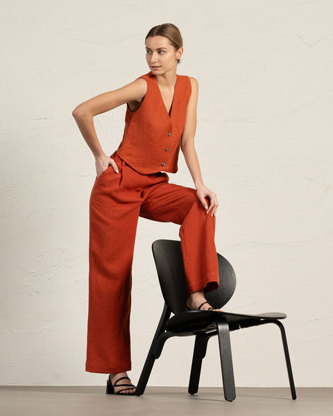 A fair-skinned woman in a cinnamon red colored linen vest and wide-leg pants poses with one leg resting on a black chair. She stands against a textured light beige wall and wears black sandals.