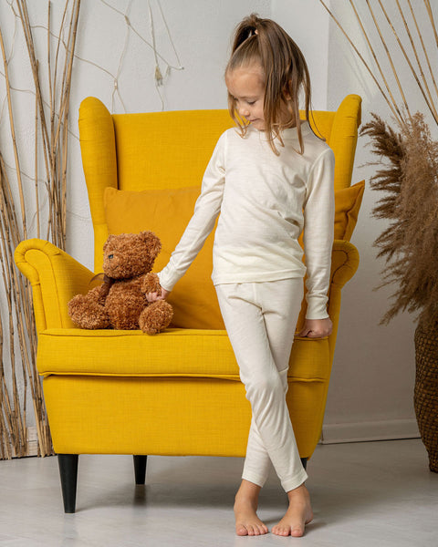 a girl standing by a yellow armchair, holding a teddy bear by the hand. She is wearing the menique merino long sleeve set in natural color.