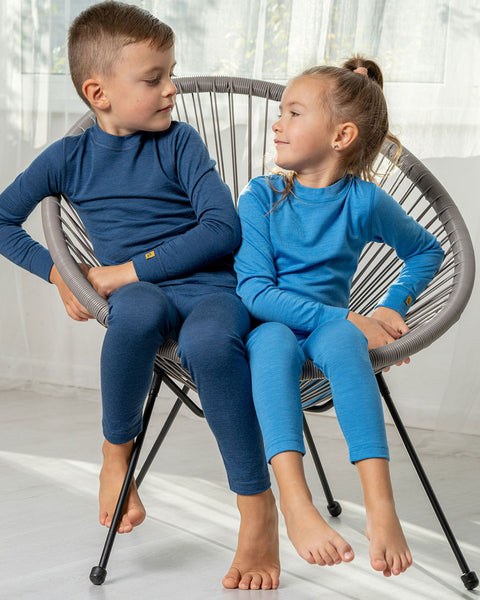Two children, a boy and a girl, wearing the menique merino 160gsm long sleeve two piece sets. The girl is wearing one in light blue color, and the boy in denim. Both of them are sitting in the same lounge chair.