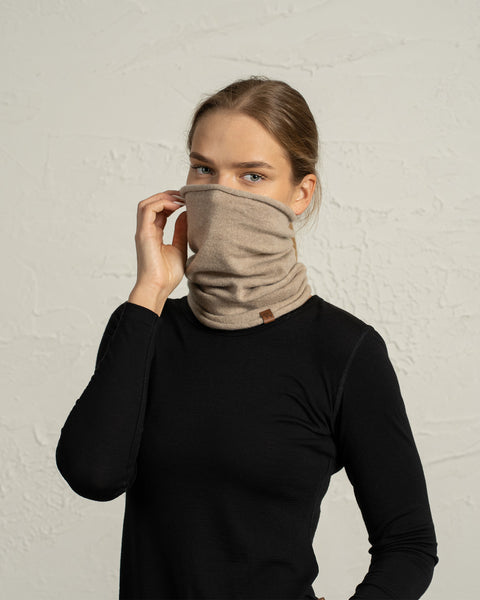 Woman adjusting a beige merino wool neck gaiter, pulling it up to cover part of her face.