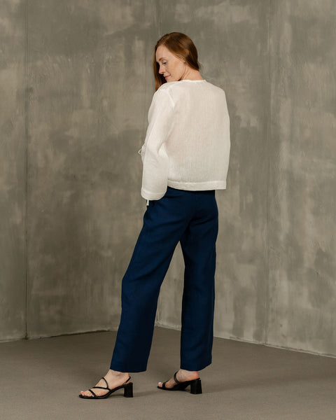 A person with long reddish-brown hair is shown from the back, modeling menique linen wide-leg trousers and a pure white long-sleeved top. They are wearing black open-toe heeled sandals and are positioned against a textured gray wall and floor.