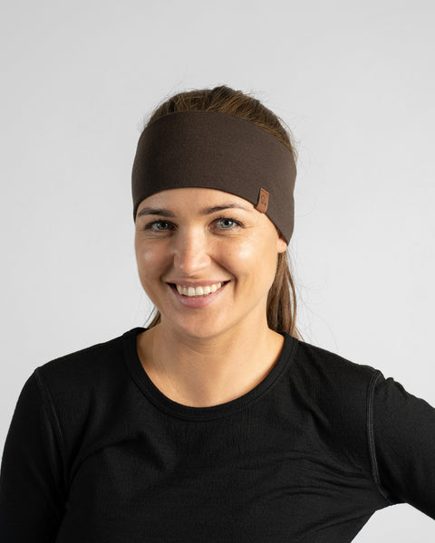 A chest-up studio portrait of a woman smiling towards the camera, wearing a dark brown knit headband and a black crewneck shirt against a neutral gray background.