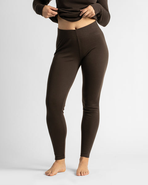A woman standing barefoot against a white backdrop, posing slightly angled to show the full front-to-side profile of a dark brown thermal base layer set.
