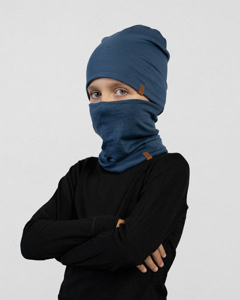A child wearing a dark blue beanie and matching neck gaiter over a black long-sleeve shirt poses with arms crossed against a light gray background.