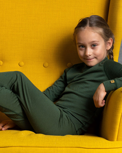 a girl, lying in a spicy yellow color ottoman, with her arm on the arm rest. She is wearing the menique Kids' Long Sleeve Crew 160 Merino in Dark Green color.