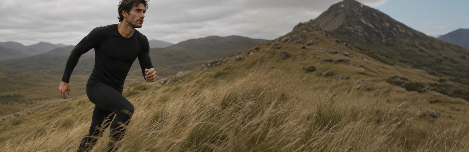 A man in a fitted black long-sleeved top and leggings runs uphill through a field of tall, dry grass. He looks determinedly ahead against a backdrop of rolling mountains and a heavily clouded sky.