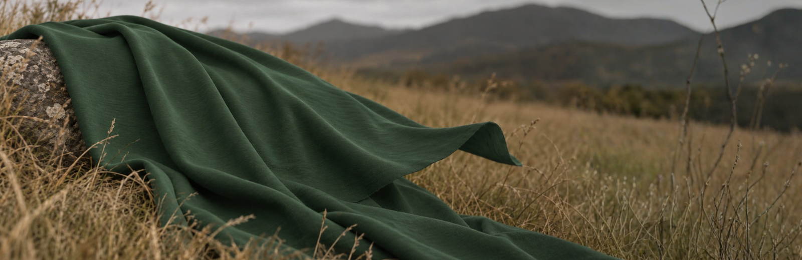 A piece of textured dark green fabric is draped over a weathered rock in a field of dry, wild grass. The fabric's edge catches a gentle breeze against a softly blurred backdrop of rolling mountains and a hazy, overcast sky.