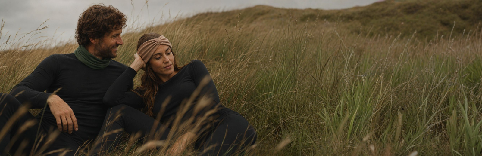 A smiling man and a woman sit closely together in a field of tall, dry grass, both wearing matching black long-sleeved tops and bottoms. The man wears a green neck gaiter while looking at the woman, who rests her hand near her tan twisted headband under a moody, heavily overcast sky.