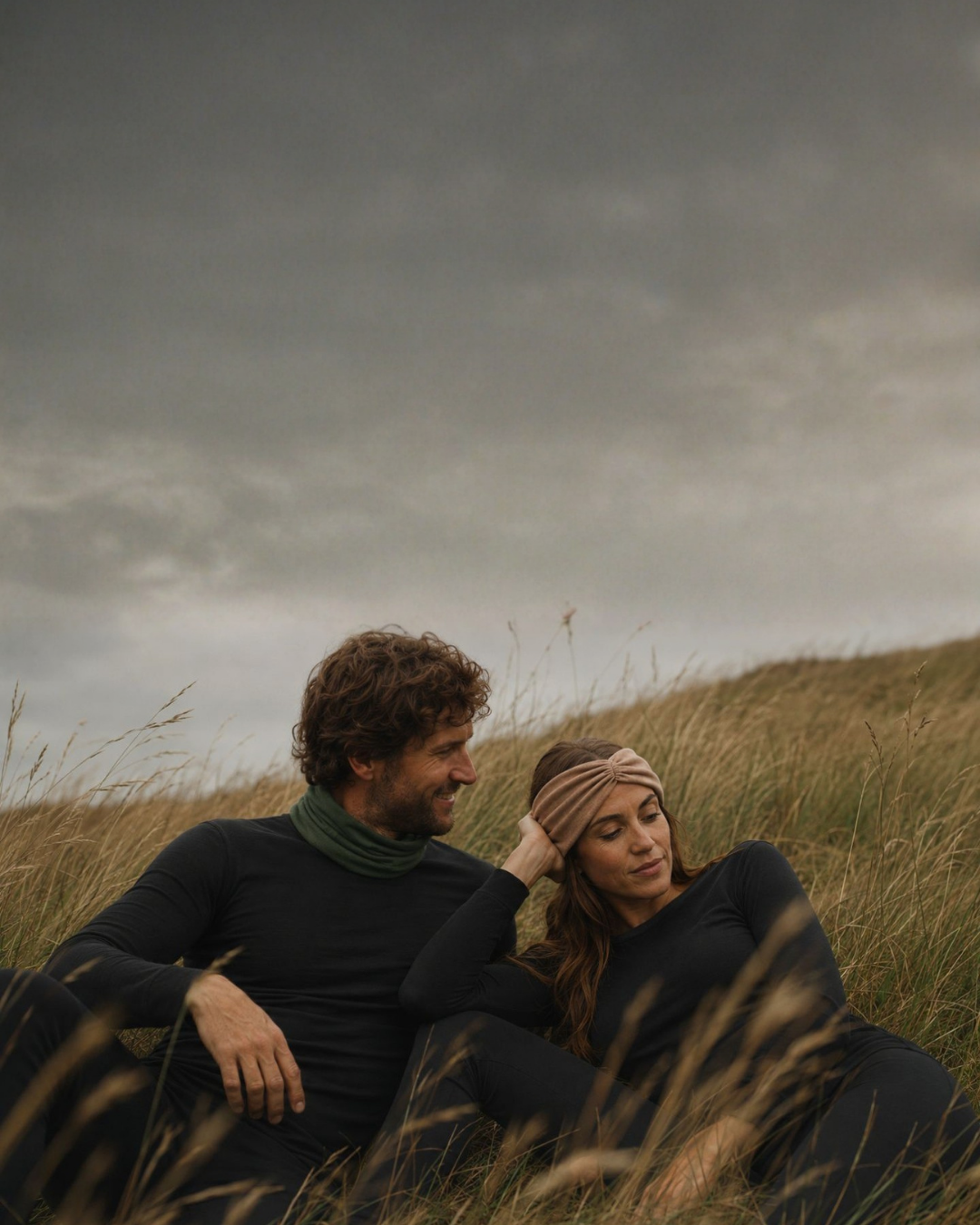 A smiling man and a woman sit closely together in a field of tall, dry grass, both wearing matching black long-sleeved tops and bottoms. The man wears a green neck gaiter while looking at the woman, who rests her hand near her tan twisted headband under a moody, heavily overcast sky.
