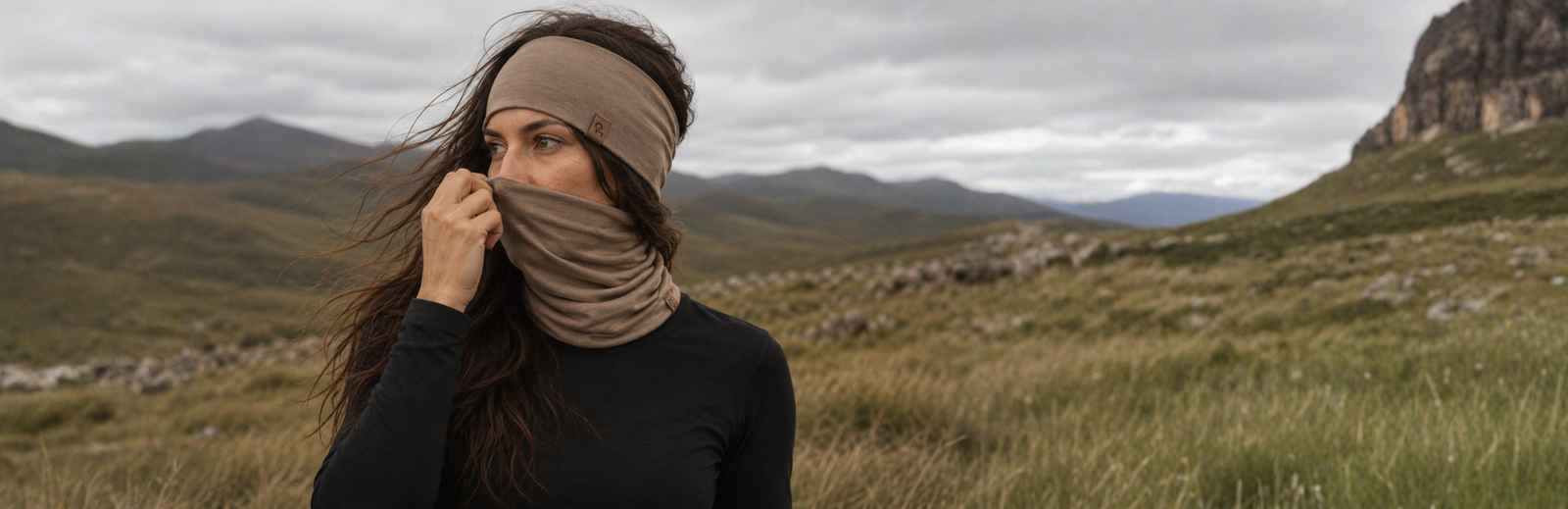 A woman with long, windblown hair stands in a rugged mountainous landscape under an overcast sky. She wears a black long-sleeved top and pulls a tan neck gaiter over her lower face to match her knit headband.