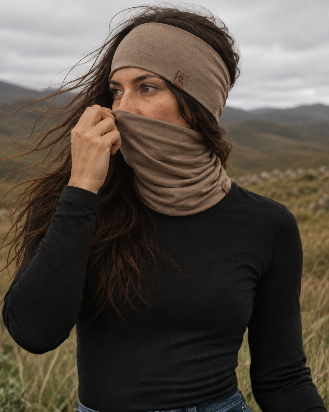 A woman with long, windblown hair stands in a rugged mountainous landscape under an overcast sky. She wears a black long-sleeved top and pulls a tan neck gaiter over her lower face to match her knit headband.