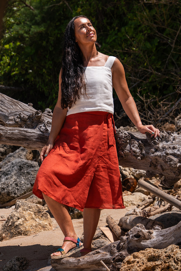 A woman wearing a white top and an cinnamon red skirt stands on a sandy area with rocks and driftwood, looking up with a smile.