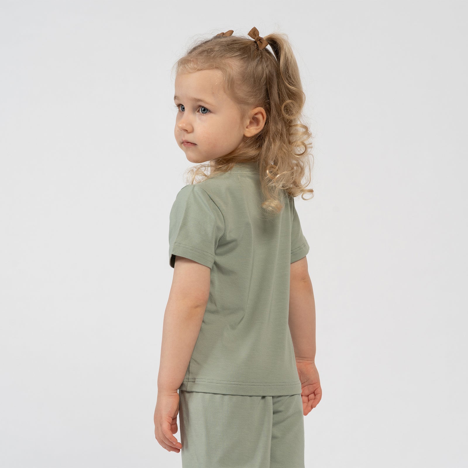 A studio portrait of a smiling young blonde girl sitting barefoot on the floor. She is angled slightly to the left, looking directly at the camera with her knees pulled up towards her chest and her hands holding her toes. She is wearing a matching set of a light sage green short-sleeved t-shirt and shorts against a plain light grey background.