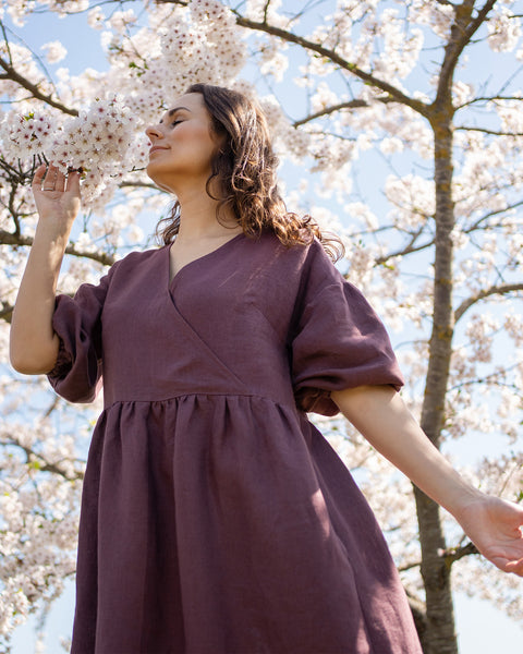 A person with dark, wavy hair and eyes closed stands outdoors, smelling the white blossoms of a tree. They are wearing a long,  shadow purple V-neck dress with loose, elbow-length sleeves. The branches of the blooming tree are visible behind them against a bright blue sky.