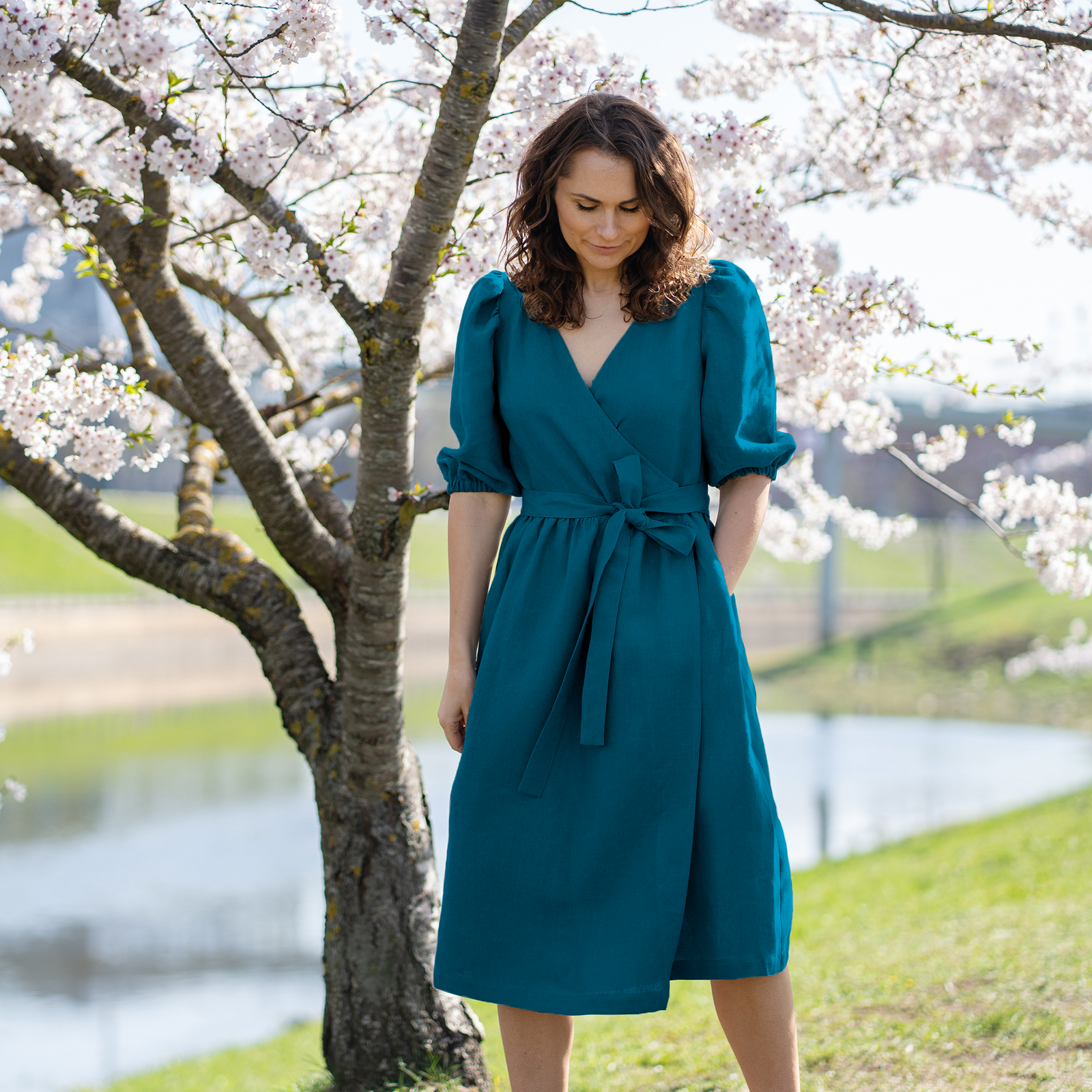 A woman with brown hair, wearing a mint green linen wrap dress with elbow-length puff sleeves, stands outdoors next to a tree with white blossoms.