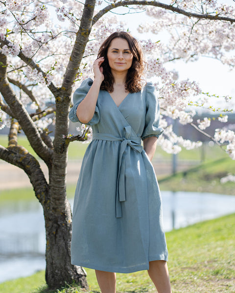 A woman with brown hair, wearing a mint green linen wrap dress with elbow-length puff sleeves, stands outdoors next to a tree with white blossoms.