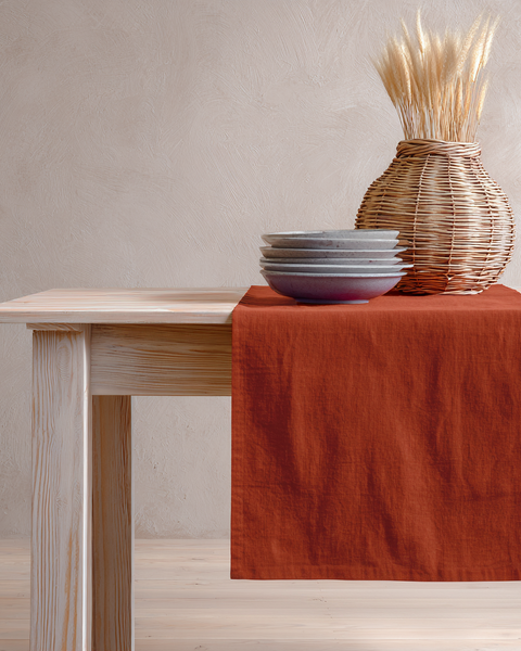 Cinnamon red linen table runner on rustic wood table with ceramic bowls and wicker vase decor.