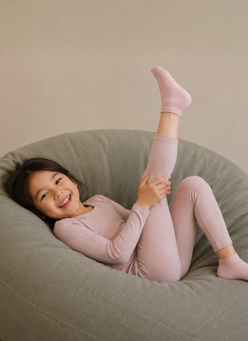 A young girl smiling while lying on a large gray beanbag chair, wearing a matching light pink long-sleeve top and leggings set with pink socks.