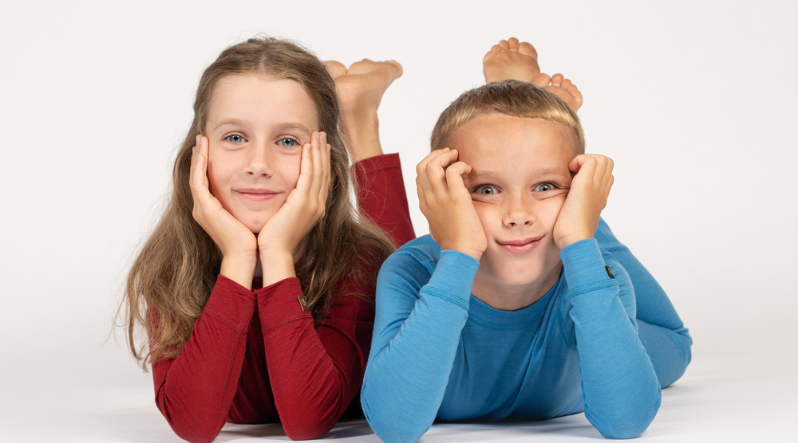 Two kids lying on floor wearing colorful long-sleeve tops, smiling at camera