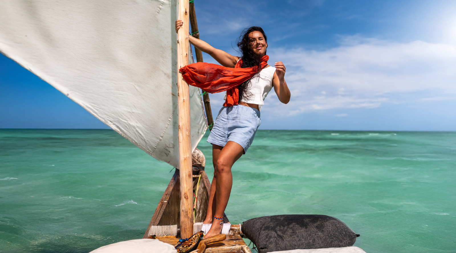 A woman stands confidently on a wooden sailboat navigating bright turquoise waters under a sunny blue sky. She wears a white top and light blue shorts, holding onto the rigging while a vibrant orange scarf catches the ocean breeze.