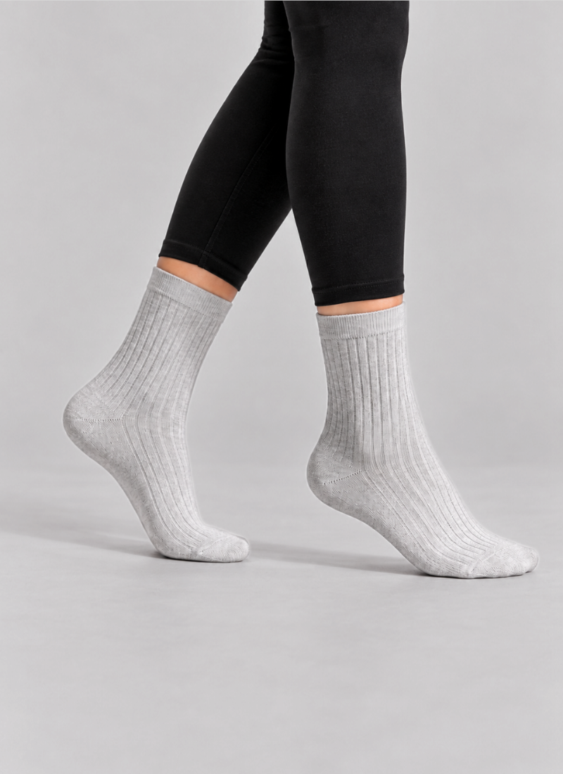 The cropped image focuses on a person's lower legs and feet as they take a mid-air step against a plain light grey studio background. They are wearing black leggings seamlessly paired with snug, light grey ribbed crew socks.