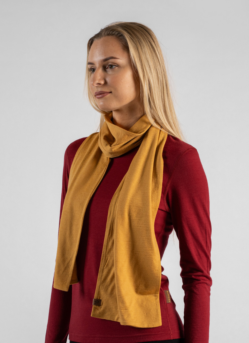 A blonde woman stands against a seamless light grey studio background, looking slightly off-camera with a neutral expression. She is wearing a dark red long-sleeved top with a mustard yellow knit scarf draped casually around her neck.