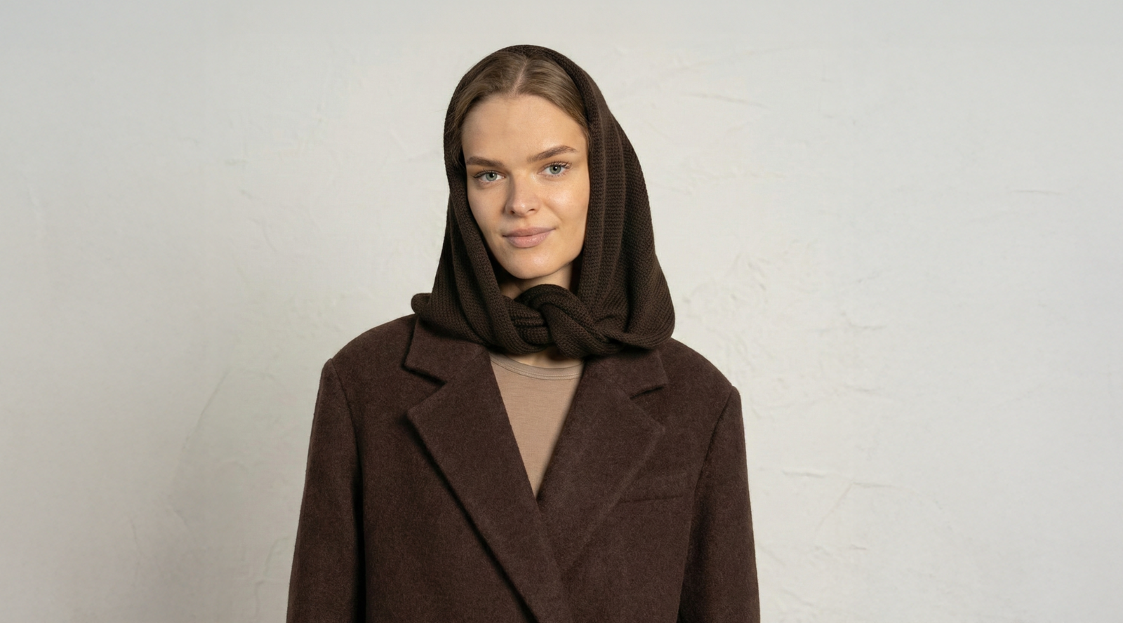 A blonde woman wearing a heavy dark brown coat over a tan top smiles softly in front of a textured off-white wall. A dark brown knit hood covers her head and wraps comfortably around her neck.