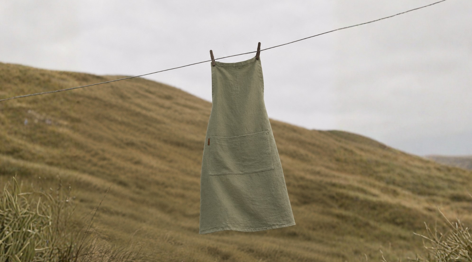 A sage green linen apron with front pockets hanging from a clothesline outdoors, set against a blurred grassy hillside.