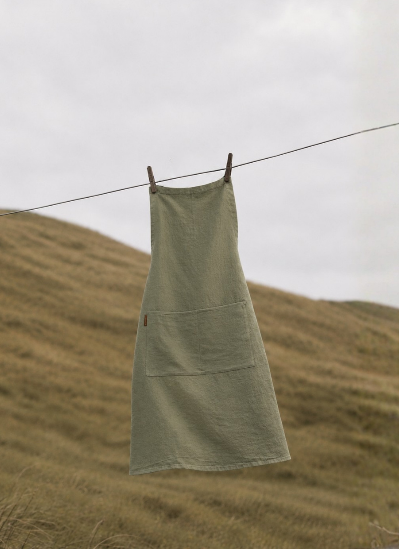 A sage green linen apron with front pockets hanging from a clothesline outdoors, set against a blurred grassy hillside.