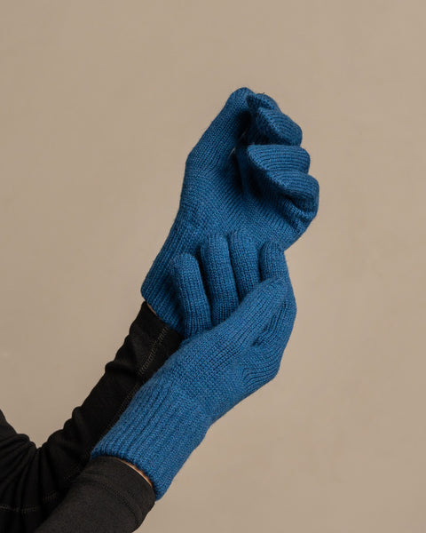 
A close-up shot of a person's hands wearing menique brand vibrant blue, ribbed knit gloves. The left hand is clenched into a fist, while the right hand appears to be gripping the wrist area of the left glove. The person is wearing a dark long-sleeved shirt underneath. The background is a solid, neutral gray.