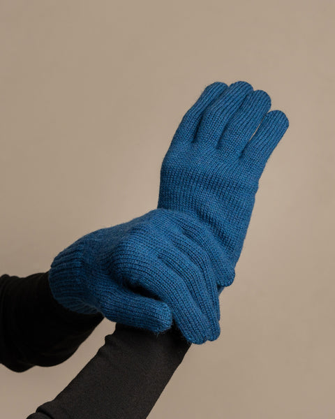 A close-up shot of a person's hands wearing menique brand vibrant blue, ribbed knit gloves. The right hand is gently supporting the underside of the left hand, which is slightly cupped. The person is wearing a dark long-sleeved shirt underneath. The background is a solid, neutral gray.