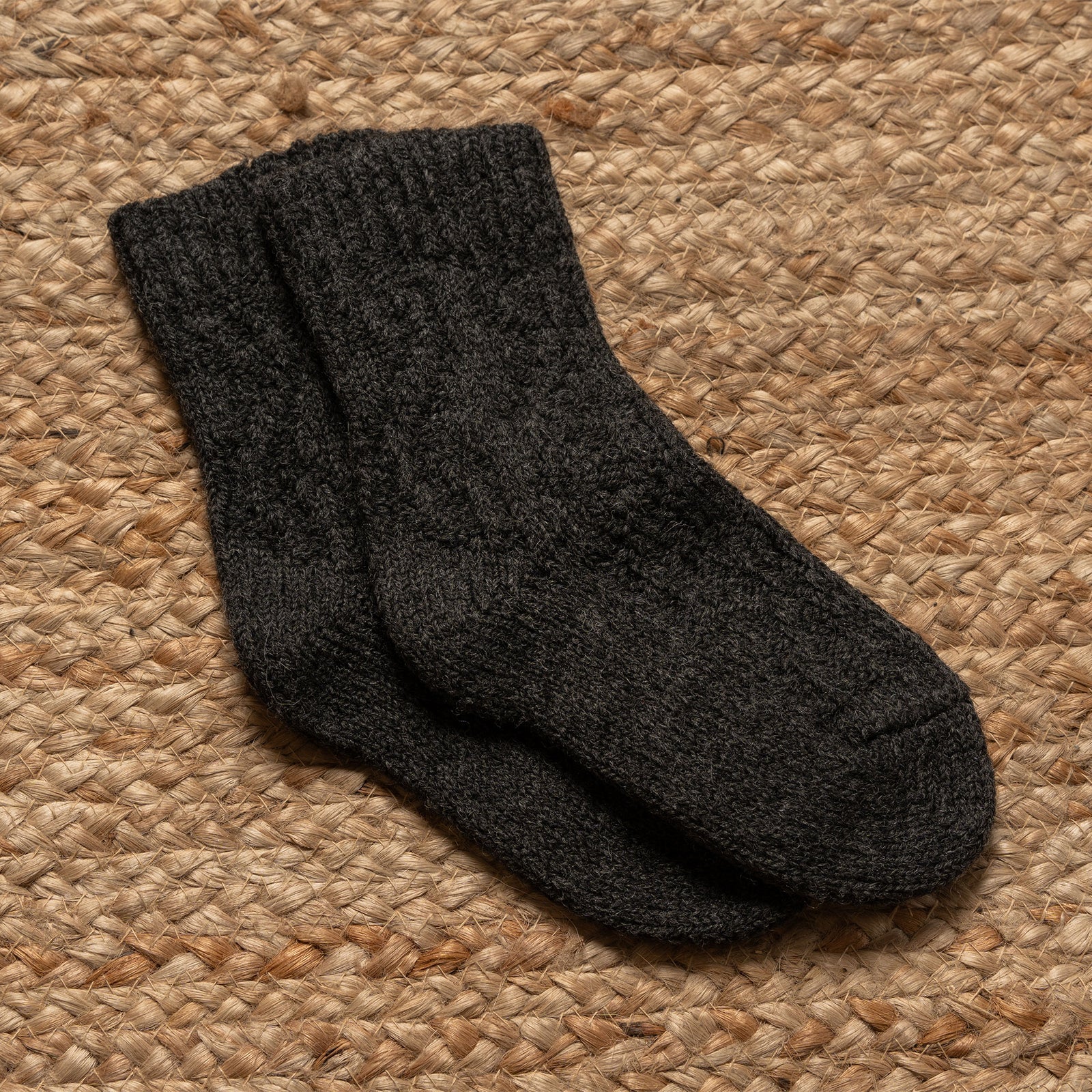 A low-angle shot shows a person's legs and feet, likely wearing menique brand off-white, crew-length knitted wool socks with a visible ribbed cuff and textured cable-knit pattern on the foot. The person is standing on a light brown, woven mat, against a neutral, plain background.